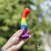 A hand holds the Avant P1 Silicone Pride Freedom Dildo, crafted from body-safe silicone in rainbow colors, against a blurred outdoor greenery background. "blush" appears in white text at the bottom right corner.